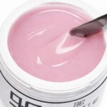Pink gel nail polish in a white container, with a brush resting on the surface and swirling the pink liquid.