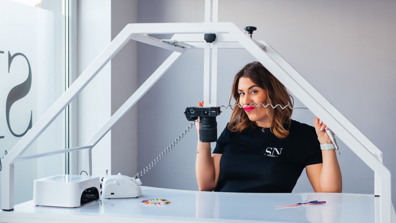 Woman in a black shirt sits at a white photography desk, holding a camera with a coiled cord in a bright studio frame structure around her.