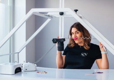 Woman in a black shirt sits at a white photography desk, holding a camera with a coiled cord in a bright studio frame structure around her.