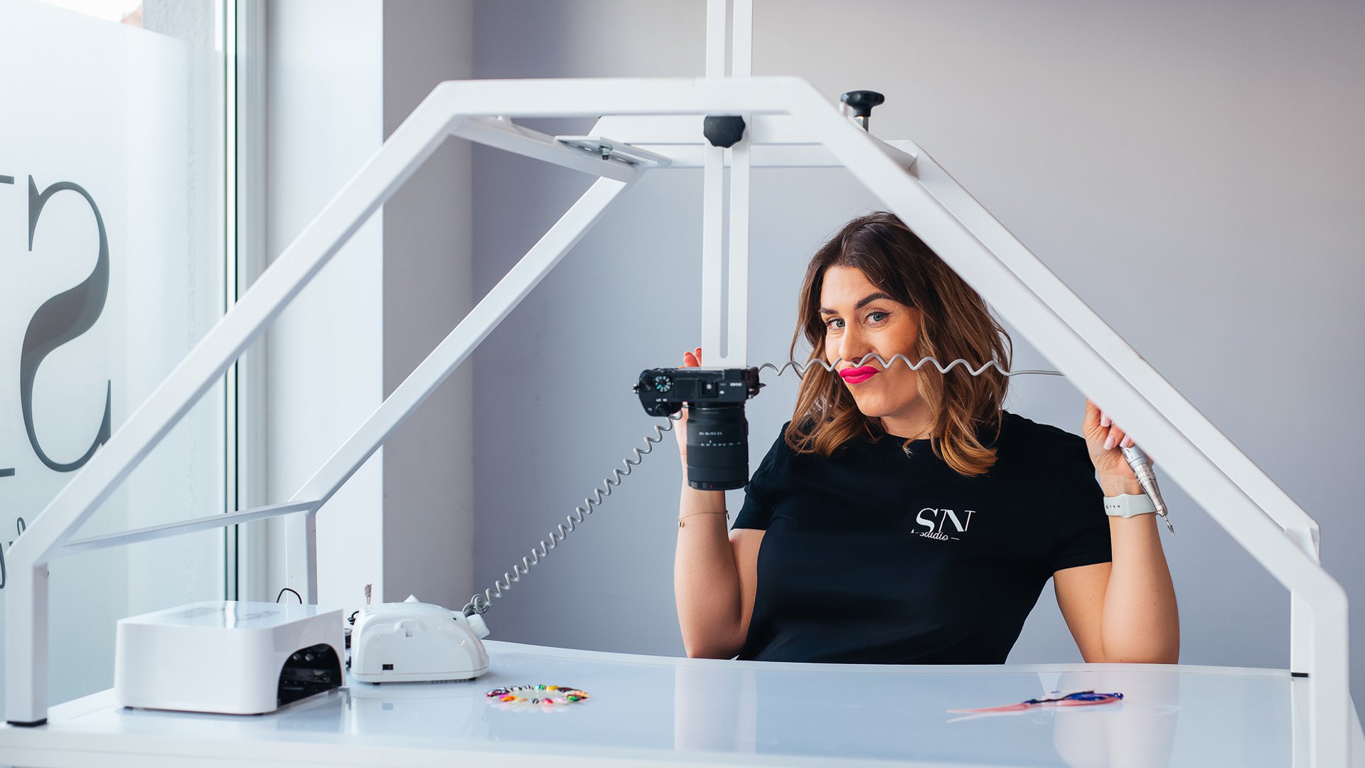 Woman in a black shirt sits at a white photography desk, holding a camera with a coiled cord in a bright studio frame structure around her.