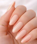 Close-up of a hand with nude glossy almond nails and a thin gold ring on the index finger.
