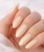 Close-up of a hand with almond-shaped nude manicure; pale cream nails and a visible gold ring on the finger.