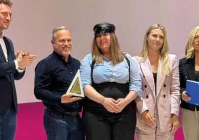 Five people stand on a stage posing for a photo; a man on the left claps while another man holds a triangular award, with three women beside them.