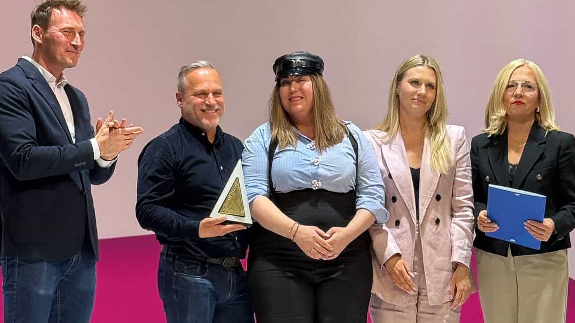 Five people stand on a stage posing for a photo; a man on the left claps while another man holds a triangular award, with three women beside them.
