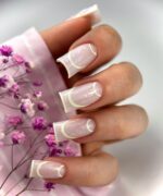 Close-up of a hand with square pale pink nails featuring white curved tips and subtle lines, held near pink flowers.